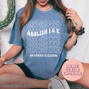 May include: Blue jean colored t-shirt with the words "ABOLISH I.C.E." repeated in a wavy font. The phrase "NO HUMAN IS ILLEGAL" is printed below. The shirt is a solid color with a crew neck.