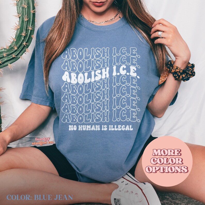 May include: Blue jean colored t-shirt with the words "ABOLISH I.C.E." repeated in a wavy font. The phrase "NO HUMAN IS ILLEGAL" is printed below. The shirt is a solid color with a crew neck.