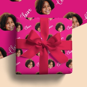 May include: A wrapped gift with a bright pink background featuring a repeating pattern of a smiling child's face and the name "Claire" in white script. A large, vibrant red bow adorns the top of the gift.