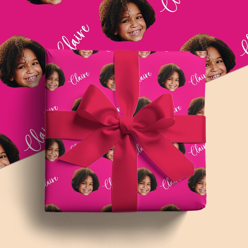 May include: A wrapped gift with a bright pink background featuring a repeating pattern of a smiling child's face and the name "Claire" in white script. A large, vibrant red bow adorns the top of the gift.