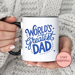 May include: White ceramic mug with a black handle and rim. The mug features the text "World's Greatest DAD" in blue script. The mug is held by a person wearing a gray sweater.