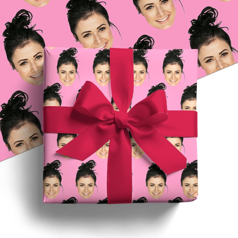 May include: A wrapped gift box with a bright pink bow. The wrapping paper is pink and covered with smiling faces. The bow is a deep pink color. The gift box is on a white surface.