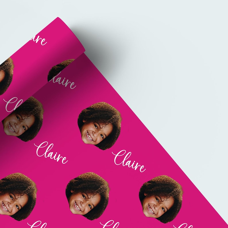 May include: Bright pink wrapping paper featuring a repeating pattern of a smiling girl's face and the name "Claire" in white cursive. The paper is partially rolled, revealing the pattern.
