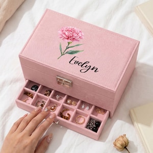 Pode incluir: Uma caixa de joias retangular rosa com tampa articulada e gaveta deslizante. A tampa apresenta uma flor de cravo rosa impressa e o nome Evelyn em letra cursiva preta. O interior é dividido em pequenos compartimentos com anéis.