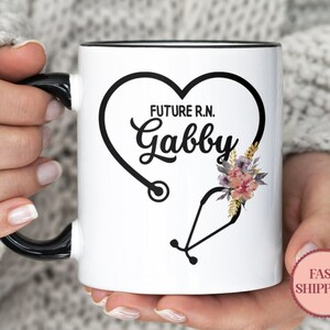 May include: White ceramic mug with a black handle and rim. The mug features a black heart-shaped design with a stethoscope, the text "FUTURE R.N. Gabby", and floral accents. A pink "FAST SHIPPING" sticker is visible.