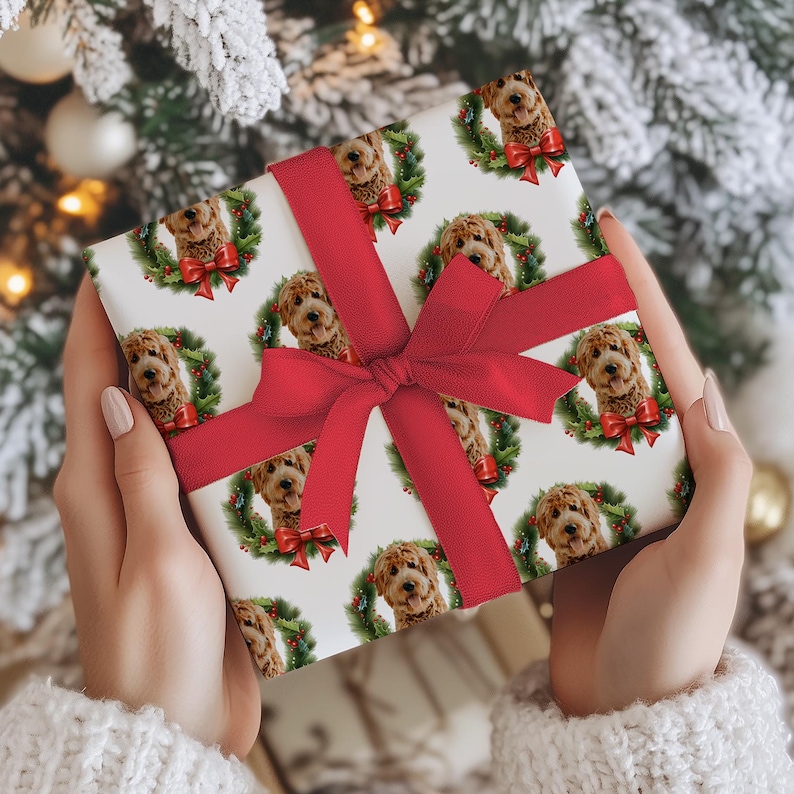 May include: A wrapped gift featuring a repeating pattern of a dog's face inside a Christmas wreath with a red bow. The gift is wrapped in white paper and tied with a red ribbon and bow. The background is a Christmas tree.