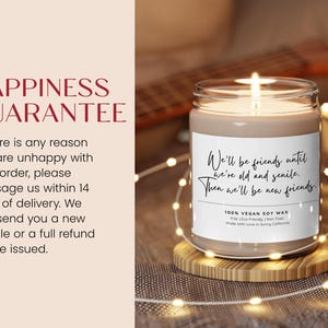 a candle on a wooden stand with a string of lights around it. The candle has a label that reads "Happiness Guarantee" and "We'll be friends until we're old and sick. Then we'll be new friends." The candle is placed on a table with a blurred background.