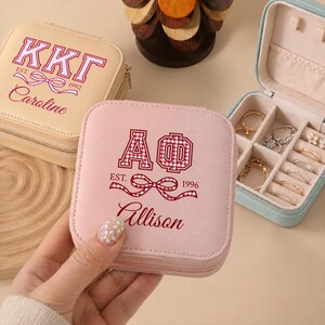 Pode incluir: Três porta-joias quadrados em bege, rosa claro e azul claro. O porta-joias bege apresenta o texto KKG EST. 1992 e Caroline. O rosa exibe AΦ EST. 1996 e Allison com um laço xadrez vermelho. O porta-joias azul está aberto, revelando vários anéis.