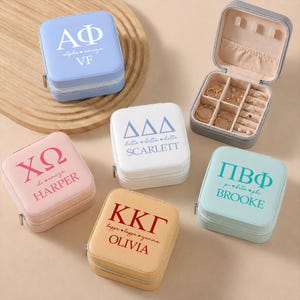 May include: Five square jewelry boxes in light blue, white, pink, tan, and mint green, each featuring Greek letters, names, and small heart icons. One box is open, revealing a beige interior with compartments for rings and small accessories.
