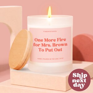 May include: A lit white candle with a wooden lid, featuring a pink label with the text "One More Fire for Mrs. Brown To Put Out." The candle is hand-poured in the USA and is 8 oz. The background is a soft pink.