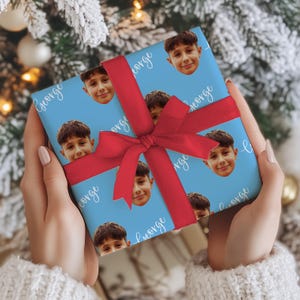 May include: A wrapped gift with a light blue background featuring a repeating pattern of a boy's face and the name "George" in white script. The gift is tied with a bright red ribbon and bow.