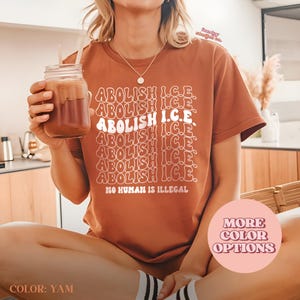May include: A brown t-shirt with the text "ABOLISH I.C.E." repeated multiple times in a stacked design. The phrase "NO HUMAN IS ILLEGAL" is printed below. A glass of a brown beverage is held in the left hand. The shirt color is "YAM".