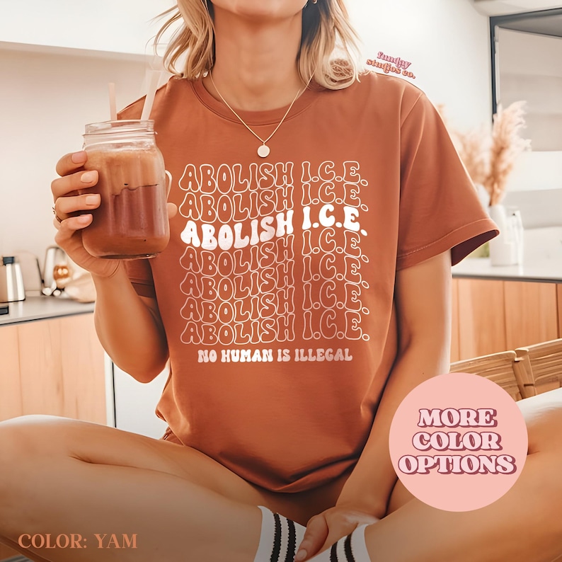May include: A brown t-shirt with the text "ABOLISH I.C.E." repeated multiple times in a stacked design. The phrase "NO HUMAN IS ILLEGAL" is printed below. A glass of a brown beverage is held in the left hand. The shirt color is "YAM".