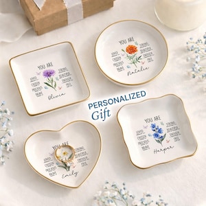 May include: Four white ceramic ring dishes with gold rims, each featuring floral designs and the words "YOU ARE" with positive affirmations. The dishes are personalized with names like Olivia, Natalie, Emily, and Harper. The words "PERSONALIZED GIFT" are also visible.