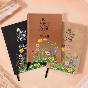 May include: Three rectangular journals in black, tan, and light brown, each featuring the text He restores my soul Psalm 23:3, a personalized name, and a colorful printed illustration of various wildflowers at the bottom with a black ribbon bookmark.