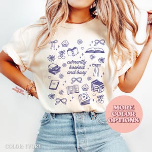 May include: Ivory t-shirt with a blue graphic design of books, bows, cupcakes, and the text "currently booked and busy." The design includes hearts, flowers, and cherries. The shirt is a casual style, suitable for everyday wear.