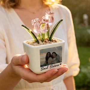 May include: A square white ceramic planter containing three translucent pink glass tulips with green glass stems and leaves, set in small light-colored pebbles. The front of the planter features a printed photograph of two people and the text Soul Sisters.