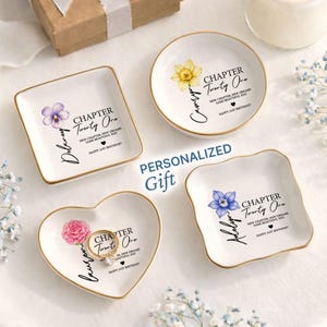 May include: Four white ceramic trinket dishes with gold-painted rims, featuring floral illustrations and black text reading "CHAPTER Twenty One," "NEW CHAPTER, NEW DREAMS SAME BEAUTIFUL YOU," and "HAPPY 21ST BIRTHDAY," with personalized names and a gold ring.