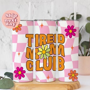 May include: Three stainless steel tumblers with pink and white checkered backgrounds and orange text that reads "TIRED MOM CLUB". Each tumbler has a daisy flower design and a clear straw.