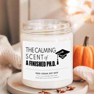 a white candle labeled "The calming scent of a finished PhD" on a white surface, with a small pumpkin and star anise nearby.