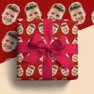 Puede incluir: Una caja de regalo envuelta en papel rojo con un patrón de caras sonrientes. Una cinta y un lazo grandes y de color rojo brillante están atados alrededor de la caja. El fondo es de color rojo liso.