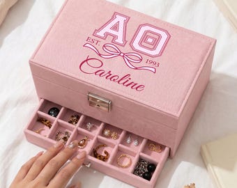 May include: A rectangular light pink jewelry box with a velvet-like texture, featuring a silver metal latch and a pull-out drawer. The lid is printed with the letters AO, a bow graphic, the text EST. 1993, and the name Caroline in script.