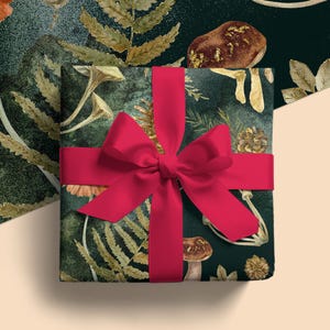 Op de afbeelding: Een vierkante geschenkdoos verpakt in donkergroen papier met een botanisch patroon van goudkleurige varens, dennenappels en paddenstoelen, gestrikt met een breed, felrood satijnen lint in een grote strik bovenop de doos.