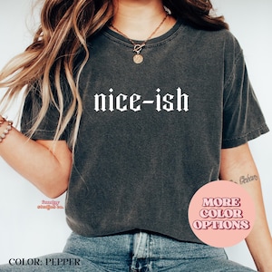 May include: A dark grey t-shirt with the words "nice-ish" in a gothic font. The shirt has a crew neck and short sleeves. A circular graphic with "MORE COLOR OPTIONS" is visible. The shirt is part of a clothing collection.
