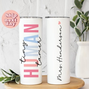 May include: Two white stainless steel tumblers with lids and straws. One tumbler has the text "tiny human" written in pink, blue, and white. The other tumbler has the text "Mrs. Henderson" written in black.