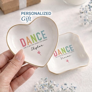 May include: Two white ceramic trinket dishes, one heart-shaped and one square, with gold rims. The heart-shaped dish is held by a hand. Both dishes have the word "DANCE" in colorful letters, the text "LIFE IS BETTER WHEN YOU DANCE", and a dancer graphic.