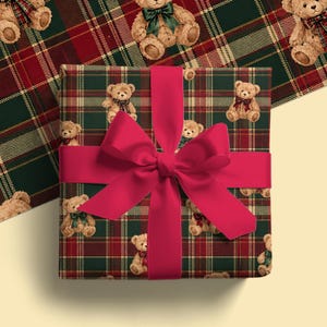 May include: A square gift box wrapped in dark green and red plaid paper featuring a repeating pattern of light brown teddy bears wearing small bows. A wide, solid bright red ribbon is tied in a large, centered bow across the front.