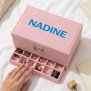 May include: A rectangular light pink jewelry box with a soft-touch finish and a silver-toned metal latch. The lid features the name NADINE printed in bold blue capital letters. A pull-out drawer reveals multiple compartments containing various rings.