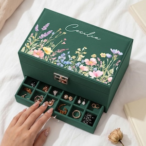 May include: A dark green jewelry box with a floral print and the name Cecilia written in white script on the lid. The box features a silver metal latch and a pull-out drawer containing multiple small compartments filled with various rings and earrings.