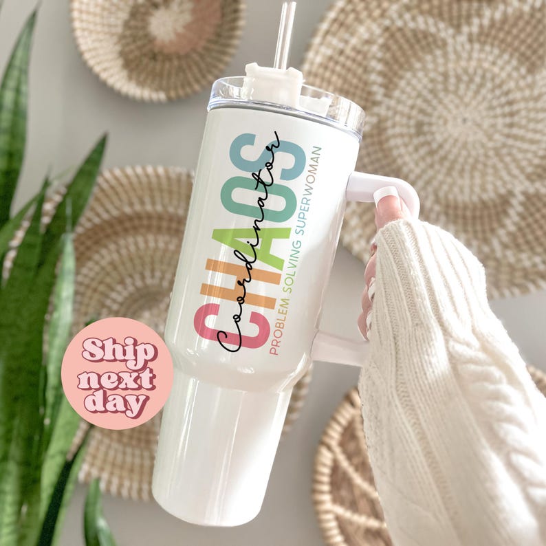 Puede incluir: Vaso blanco con asa y tapa transparente. El vaso tiene las palabras "Chaos Coordinator Problem Solving Superwoman" en texto colorido. Una pajita transparente est&aacute; insertada en la tapa. El vaso lo sostiene una persona que lleva un su&eacute;ter blanco.