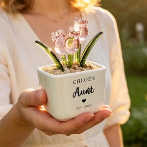 May include: A square white ceramic planter containing three translucent pink glass tulips with green glass stems and leaves, set in small beige pebbles. The front of the planter features the black text "CHLOE'S Aunt" above a small heart and "EST. 2026".