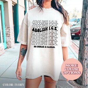 May include: Ivory-colored t-shirt with the text "ABOLISH I.C.E." repeated multiple times in a stacked design. The phrase "NO HUMAN IS ILLEGAL" is printed below the stacked text. The shirt has short sleeves and a relaxed fit.