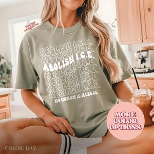 May include: Sage green t-shirt with the text "ABOLISH I.C.E." repeated multiple times in white, along with the phrase "NO HUMAN IS ILLEGAL." The shirt is a relaxed fit and has short sleeves. The color is "BAY".