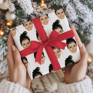 May include: A wrapped gift with a white background covered in smiling faces. The gift is wrapped with a red bow. The gift is held by a person wearing a white sweater.