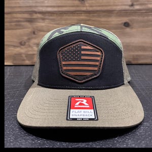 May include: A flat bill snapback hat with a camouflage top, black front panel, and a brown and black American flag patch. The hat has a tan brim and mesh back. The tag reads "Richardson Flat Bill Snapback".