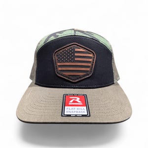 May include: A black and camouflage trucker hat featuring a brown leather-like hexagonal patch with an embossed American flag design. The flat brim includes a rectangular sticker with the text Richardson, Wear The Best, Flat Bill Snapback, Est 1970.