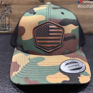 Puede incluir: Gorra de béisbol de camuflaje Woodland con parte trasera de malla negra y parche de cuero marrón con diseño de bandera americana. La gorra tiene visera curva y cierre snapback. El texto "Woodland Camo" es visible.