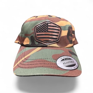 May include: A camouflage baseball cap featuring a brown and black pentagonal patch on the front with an embossed American flag design. The brim includes a circular sticker that reads The Authentic Yupoong Adjustable Yupoong Since 1974.