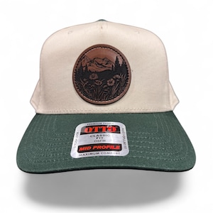 May include: A cream-colored baseball cap with a dark green brim. Centered on the front is a circular brown leather patch featuring an embossed mountain and forest landscape with wildflowers. A sticker on the brim reads OTTO CLASSIC FIT OSFM.