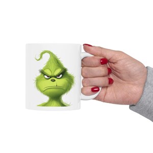 May include: A white ceramic mug featuring a graphic illustration of a green, furry creature with a tuft of hair, furrowed eyebrows, and a stern, frowning expression. The character has yellow eyes and a small nose, centered on the mug.