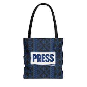 Puede incluir: Un bolso tote negro con un patrón geométrico azul y negro. La bolsa presenta un rectángulo blanco con la palabra "PRESS" en letras azules en negrita. La bolsa tiene asas negras.