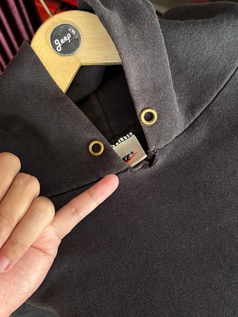 May include: Close-up of a dark gray hoodie with a drawstring hood and gold-colored eyelets. The Nike brand label is visible at the neckline. The hoodie is hanging on a wooden hanger.