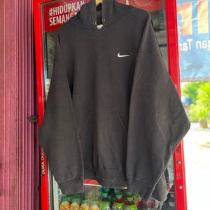May include: A dark gray Nike hoodie with a white Nike swoosh logo on the chest. The hoodie has a drawstring hood and a front pocket. The hoodie is hanging on a wooden hanger in front of a red vending machine.