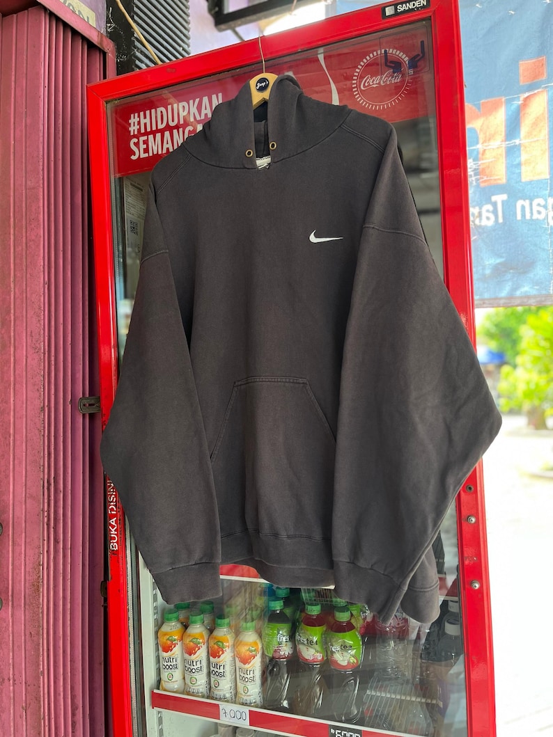May include: A dark gray Nike hoodie with a white Nike swoosh logo on the chest. The hoodie has a drawstring hood and a front pocket. The hoodie is hanging on a wooden hanger in front of a red vending machine.