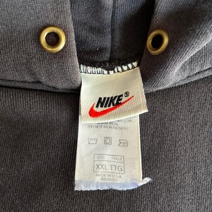 May include: Close-up of a dark gray Nike hoodie with a hood and drawstring eyelets. The Nike logo is visible on a tag, along with size information and care instructions. The tag reads "XXL TTG" and "MADE IN U.S.A."
