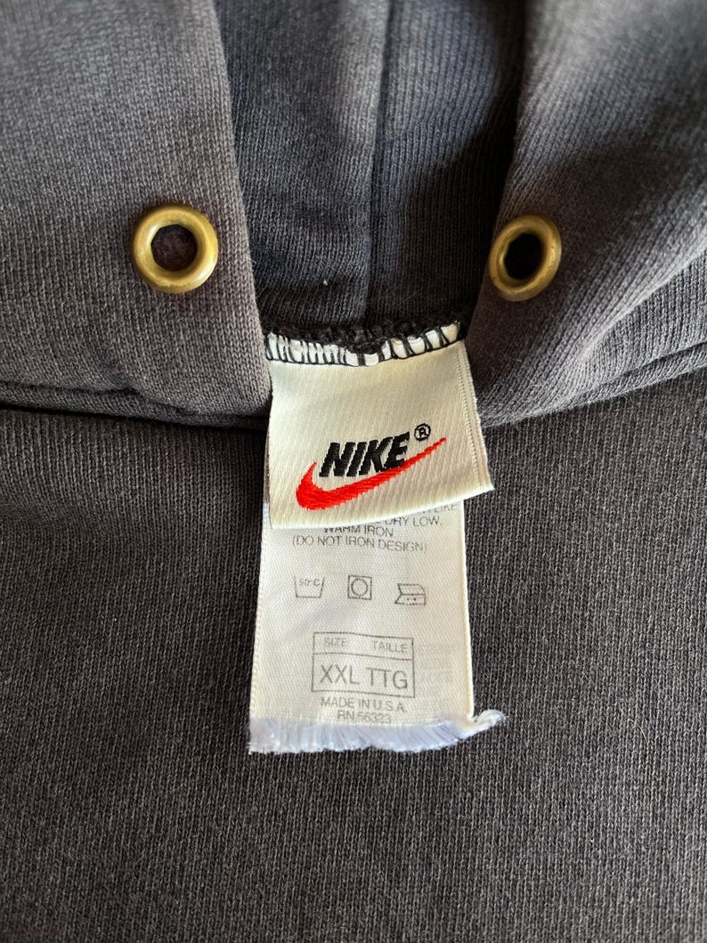 May include: Close-up of a dark gray Nike hoodie with a hood and drawstring eyelets. The Nike logo is visible on a tag, along with size information and care instructions. The tag reads "XXL TTG" and "MADE IN U.S.A."